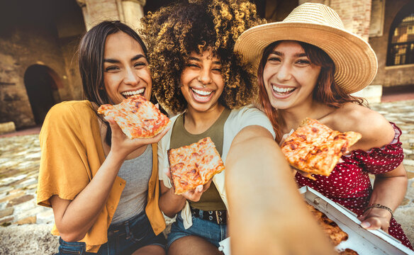 Fototapeta Three multiracial women eating pizza in city street - Smiling female friends taking selfie enjoying summer vacation in Italy
