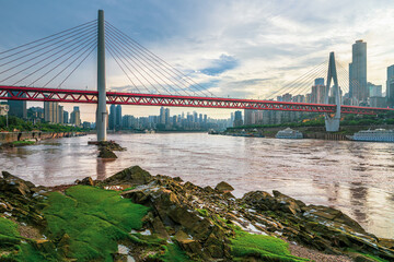 Panoramic view of a modern red suspension bridge crossing a wide river with the urban skyline of Chongqing, China.