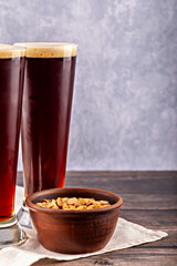 Close-up of two tall glasses of dark beer with foam next to clay bowl filled with roasted peanuts. Rustic wooden table and neutral background. Ideal for pub or bar. Two dark beers and rustic peanuts