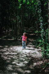 Obraz premium Smiling 7-year-old girl enjoying a forest adventure, walking among tall trees with sunlight streaming through leaves, wearing casual clothes.
