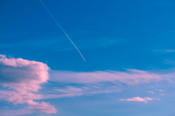 White contrail from a high-altitude airplane flying in a vibrant blue sky, with pink and white...
