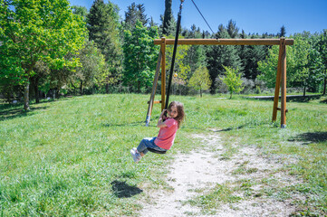 Little girl swinging on a rope in the park, wearing casual pink t-shirt and jeans, surrounded by trees, full of summer vibes.