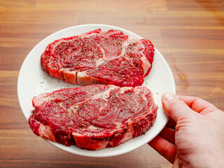 Hand holding plate with two fine raw rib eye steaks seasoned with herbs and pepper. Rich expensive beef ready to be cooked. Fine cuts with excellent marbling.