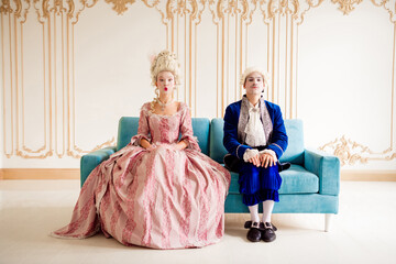Elegant couple in baroque outfits posing on a stylish sofa in a luxurious pastel-themed palace...