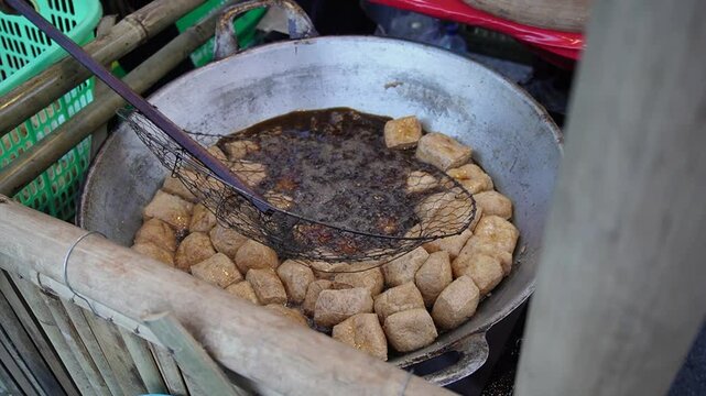 Street food vendor frying tofu or tahu goreng in bubbling hot oil at a traditional food stall. Golden tofu pieces sizzle in the pan, creating a crispy texture and savory aroma