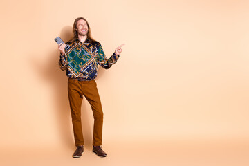 Stylish young man with long hair gesturing happily while holding a smartphone, posing against a beige background