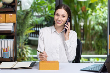 Smiling young woman talking on phone while preparing a package for delivery at her home office. Concept of online business, e-commerce, and remote work.