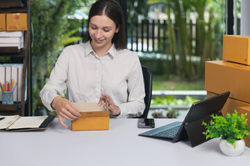 A young woman packing a cardboard box at her desk in a home office. E-commerce entrepreneur preparing orders for online shipping. Small business startup concept.