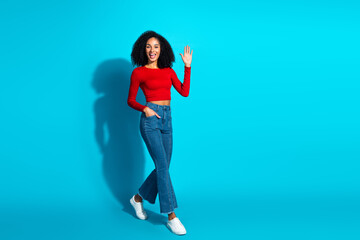 Cheerful young woman waving in casual red top and jeans against blue background, exuding happiness and style