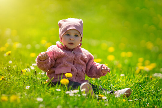 Adorable cute baby girl sitting on grass with flowers in park. Cute kid portrait. 9 month old child. Childhood concept.