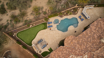 an aerial view of a desert landscaped backyard featuring a travertine tiled pool deck, putting green, outdoor kitchen and fireplace.