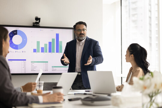 Business executive leading presentation for partners, standing in front of digital display screen showing analytics charts, engaged in investment strategy discussion, financial planning and analysis