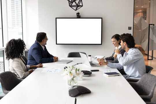 Group of four business professionals sitting around conference table in modern meeting room, looking at digital screen with white mock up, engaged in videocall, formal discussion or brainstorm event