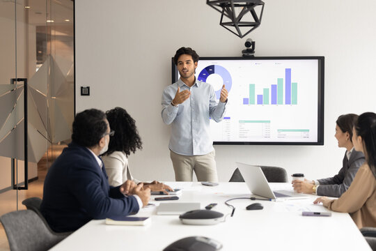 Young male professional stands in front of digital display screen presenting business data in bar and pie charts to group of colleagues seated around conference table. Strategy development, analytics - Powered by Adobe
