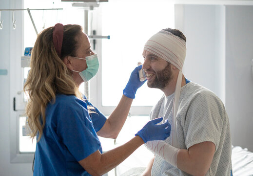 Nurse wearing surgical mask cleans face of a smiling patient with head bandages in hospital room - Powered by Adobe