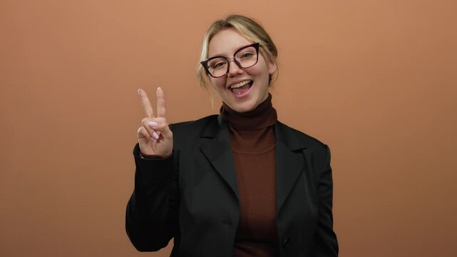 Woman smiling while making a victory sign with fingers, standing against a brown wall, wearing glasses and a blazer, showcasing a confident and cheerful expression.