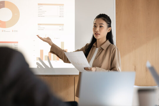 Skilled Asian business coach presenting data to colleagues during seminar or presentation in conference room, gestures to visualized charts, holds printed materials, lead analytical briefing for staff