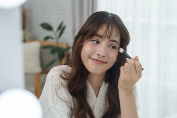 Multiracial Caucasian teenage girl looking into mirror while applying makeup using flat brush wearing white robe with lighting in room showing confidence and happiness during daily beauty care routine
