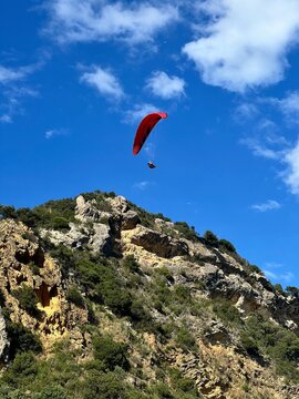 Man Flying with a red parachute, paraplane in the air. Bright, clear sky, Green mountains in the background. Boulder on a picturesque beach. Space, nature concept, retreat, sport, hobby, recreation