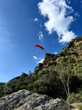Man Flying with a red parachute, paraplane in the air. Bright, clear sky, Green mountains in the background. Boulder on a picturesque beach. Space, nature concept, retreat, sport, hobby, recreation