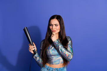 Young woman pondering over a hair iron against a vibrant purple background, wearing shiny casual attire