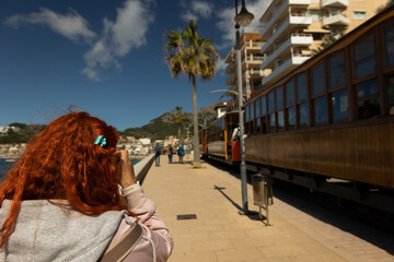 Close up of a red haired woman photographing a moving vintage tram on a sunny day next to a port