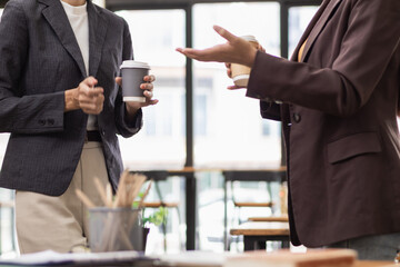 Two Asian businesswomen standing holding cups hot coffee to drink while discussing business investment together in conference room during a break. businesswomen standing holding paper cups hot coffee