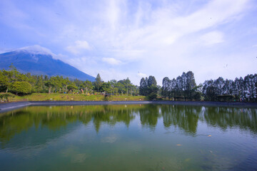 Obraz premium Serene Mountain Lake Reflection: A tranquil lake reflects the majestic silhouette of a distant mountain, framed by lush greenery under a vibrant blue sky.