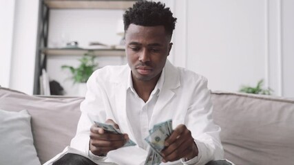 Young african american doctor counting money in his office in clinic, rich man. Black male person in white gown holding cash dollars in hands, salary and benefits for health professional, healthcare