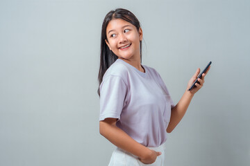 Fototapeta premium Happy young Asian woman looking away while holding a smartphone in one hand and the other in her pocket. Casual pastel outfit, smiling expression, isolated on grey background.