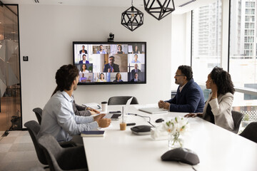 Group of multiethnic professionals sit at conference table, engaged in virtual meeting, looking at...