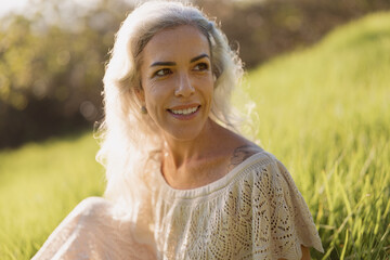 A joyful woman with grey hair smiles warmly while sitting in a grassy field. The sunlight illuminates her face, creating an atmosphere of peace and contentment.