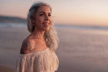 A confident mature woman with gray hair smiles serenely as she stands on the beach, enjoying a...