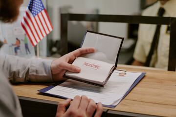 Hand holding passport stamped with rejected over desk in immigration office, with American flag in...