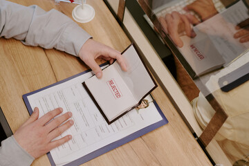 Close-up of hand holding rejected document on clipboard with paperwork on desk. Person wearing...