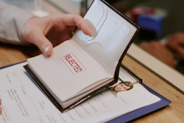 Hand opening passport displaying large red rejected stamp on page accompanied by some personal...