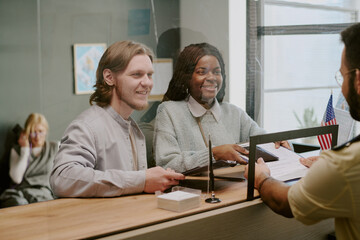 Obraz premium Smiling Caucasian man and African American woman discussing with visa officer at front desk of office building having pleasant conversation behind glass screen