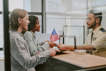 Diverse couple handing passports at immigration office counter with officer verifying documents and...