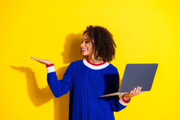 Happy young woman wearing blue knit sweater holding laptop and gesturing against a bright yellow...