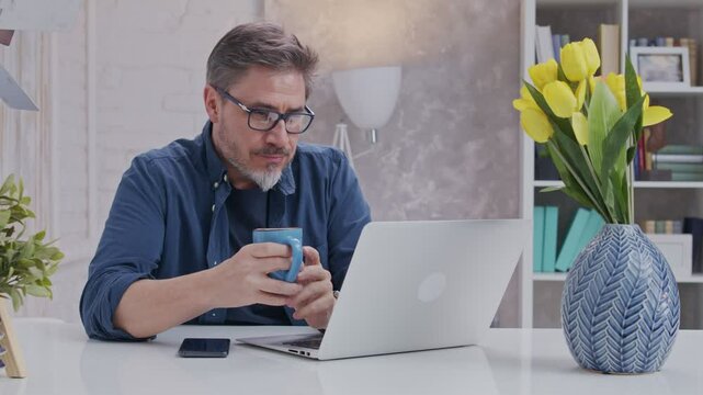 Bearded man working online with laptop computer at home sitting at desk. Home office, browsing internet, drinking coffee. Portrait of mature age, middle age, mid adult man in 50s.