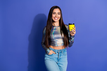 Young woman in stylish shiny top holding yellow coffee cup and smiling confidently against vibrant...