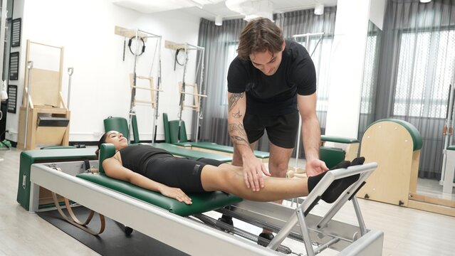 Happy pilates instructor assisting a client in performing a leg press exercise on a reformer for improving core stability. Sporty girl using pilates machine posing arm circle on arc barrel. Habituate. - Powered by Adobe