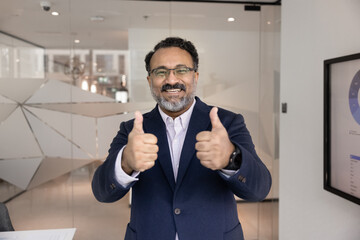 Portrait of smiling mature 45s Indian businessman in formal suit stands in office showing thumbs up celebrate great work outcome, good revenue and sales results. Business growth and financial success