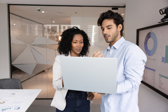 Two colleagues working in office using laptop, review presentation, check slides before meeting, analyze data, examining charts or graphs from business report, stand next to screen displaying diagrams