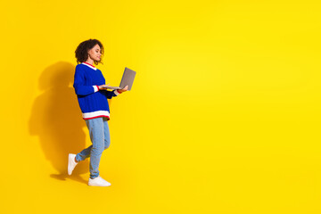 Young woman in a blue sweater using a laptop on a vibrant yellow background suggesting modern technology, casual lifestyle