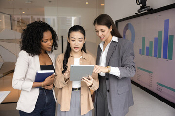 Multiethnic professional women standing in front of screen displaying analytical charts, focused on tablet, reviewing or discussing digital content, working together engaged in collaboration in office
