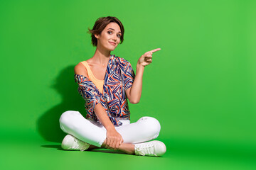 Full length photo of funny cheerful lady dressed print shirt legs crossed pointing empty space isolated green color background