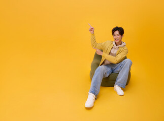 Happy young asian man sitting on bean bag pointing finger up isolated on yellow copy space...