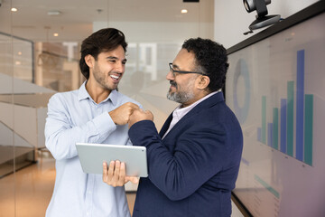 Young 30s and mature 45s multiethnic teammates standing in modern office, sharing fist bump, gesture of camaraderie, mutual respect, and celebration of joint success. Strong collaboration and teamwork