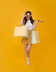 Happy celebrating cute Asian woman shopper carrying colorful shopping bags isolated on yellow background. shopping online smartphone concept.